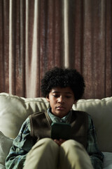 Fototapeta premium Young boy with curly hair sitting on couch in living room, concentrating on mobile phone screen, wearing casual clothing