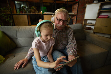 Focused child girl and elderly man sitting on couch in headphones and interacting with digital...