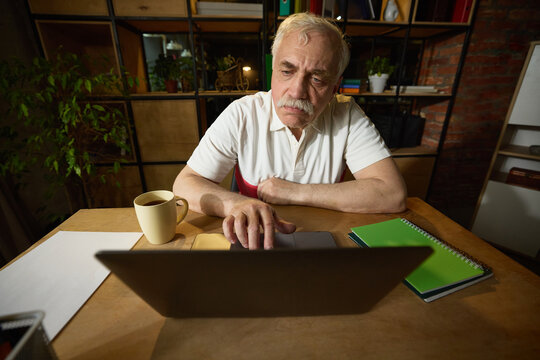 Elderly man with mustache sitting at wooden desk, looking at laptop with both hands on touchpad, mug and notebooks on the desk. Concept of elderly lifestyle, education, knowledge