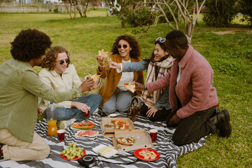 A group of five friends sit on a blanket and smile while holding slices of pizza in front of them,...