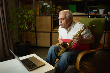 Elderly man with mustache sitting in green armchair, holding saxophone and looking at laptop,...