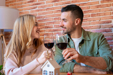 Happy caucasian couple toasting with red wine and smiling lovingly at each other. A miniature house and keys on the table symbolize the joy of moving into a new home in a cozy, modern space. 