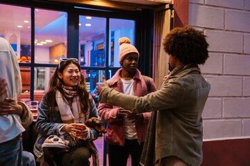 A woman sits and laughs while holding a glass and a dog while a group of four friends stand and hug next to her, outdoors