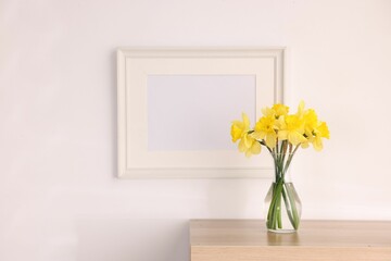 Beautiful daffodils in vase and picture frame on cabinet at home