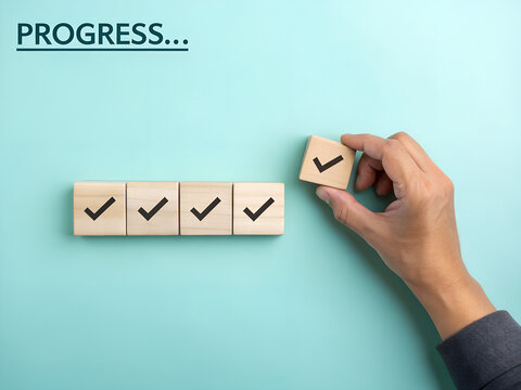Wooden blocks with check marks are arranged to show progress as a hand places a final one on a blue surface.