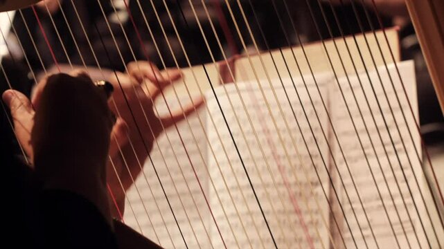 Close-up of a musician playing the harp during a classical music concert. Fingers delicately pluck the strings as sheet music rests in the background, evoking elegance, precision, and atmosphere