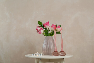 Elegant holders with burning candles and tulips in vase on white table near beige wall