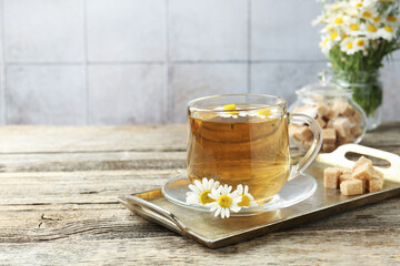 Delicious chamomile tea in glass cup, fresh flowers and brown sugar on wooden table, closeup. Space for text