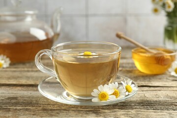 Delicious chamomile tea in glass cup, fresh flowers, honey and teapot on wooden table, closeup