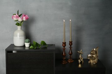 Elegant candlesticks with burning candles, tulips in vase and other decor on dresser near grey wall