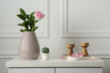 Elegant candlesticks with burning candles, tulip in vase and other decor on dresser near white wall