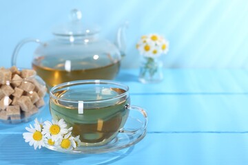 Delicious chamomile tea in glass cup, fresh flowers and brown sugar on light blue wooden table, closeup. Space for text