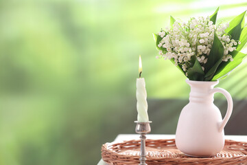 Beautiful lilies of the valley flowers in vase and candle on white table against blurred background. Space for text