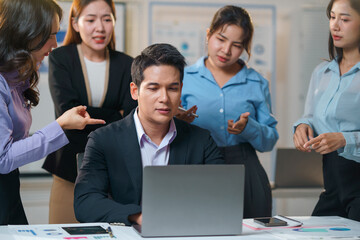 Stressed businessman working on a laptop while colleagues express blame during a tense office meeting filled with conflict and frustration