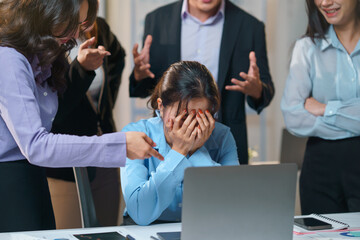 Colleagues pointing at businesswoman covering face with hands while sitting at desk in office...