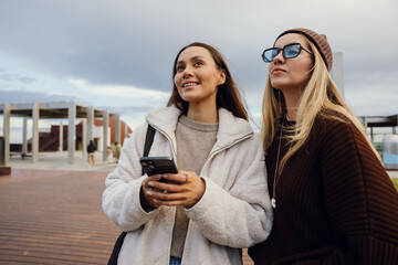 Plakat Two female friends standing and looking ahead while one of them smiles and holds a phone, outdoors