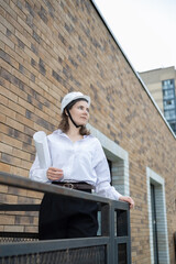 A female engineer inspector in a white helmet and white shirt.In the background there is a construction site and tower cranes In her hands drawings and papers