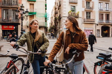 Two female friends looking at each other and holding bicycles, outdoors