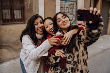Group of three female friends standing and hugging while smiling and holding bottles while looking at the phone one of them is holding, outdoors