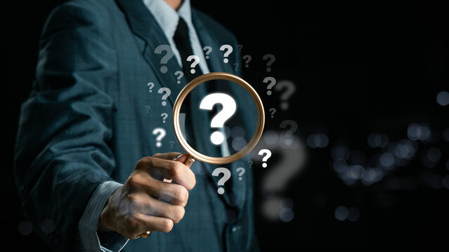 A hand holding a magnifying glass over large question marks surrounded by smaller ones symbolizes cause analysis and problem-solving. Question mark and root cause analysis concept. solution, analyze