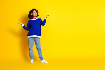 Cheerful young woman in casual blue sweater poses joyfully against vibrant yellow background demonstrating open hand gestures