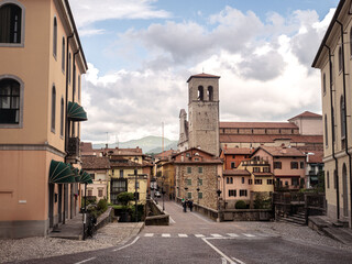 The heart of Cividale del Friuli, Italy, with colorful buildings, a cobblestone street, and the prominent bell tower rising against a backdrop of clouds and distant mountains around the Devil's Bridge