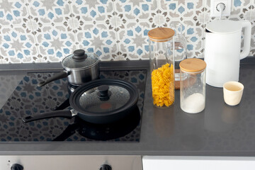 Modern minimalist kitchen with clean black countertop, electric stove, cooking pots with lids, glass storage jars filled with pasta and salt, white electric kettle, and ceramic cup
