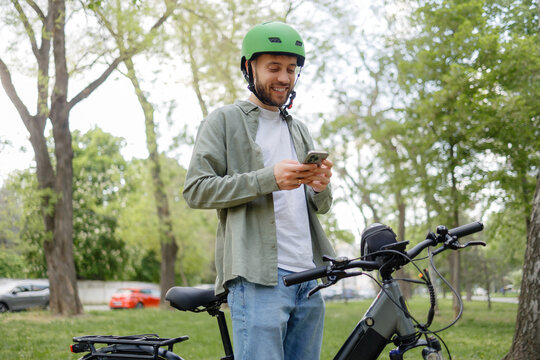 Smiling man in green helmet checks messages while standing by bicycle in a park during daytime