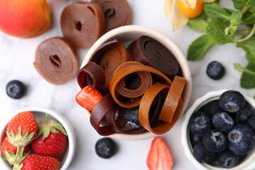 Tasty fruit leather rolls in bowl, mint and berries on white marble table, flat lay