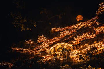 Traditional Chinese architecture glowing at night in Wangxian Valley Town with a full moon rising behind the mountain, creating a magical and serene atmosphere.