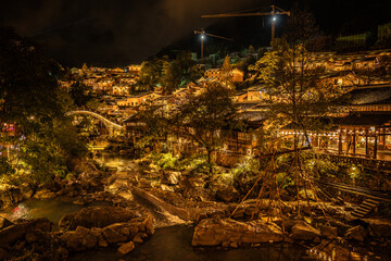 Night view of Wangxian Valley Town with a glowing arched stone bridge over a river, surrounded by traditional Chinese buildings and warm lights in the hillside town.