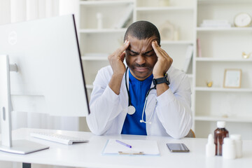 African American male doctor in white coat sitting at desk, holding his head with both hands, showing signs of stress and fatigue, medical worker suffering headache, copy space
