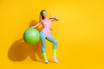 Young woman in vibrant retro activewear posing joyfully with yoga ball on yellow background,...