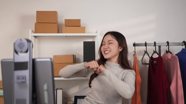 A woman is dancing in front of a livestreaming matchine live or recording video in a room with hanging clothes and pacel boxes