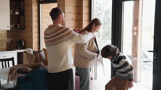 An adult man and woman with a school-aged boy indoors unpack groceries in a wooden house. The man helps the woman with her coat while the child bends down near the doorway.