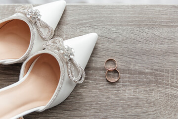 White bridal shoes decorated with rhinestones and crystals are lying near two golden wedding rings on a gray wooden background, creating a stylish and elegant wedding composition