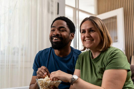 people and leisure concept - diverse couple with popcorn watching tv at home. Couple watching a movie. Relaxed couple with popcorn and remote control - Powered by Adobe