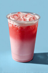 Iced strawberry milk drink with creamy foam on top in clear plastic cup on blue background, refreshing and cold