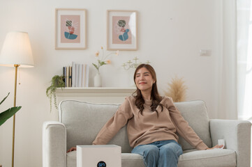 Healthy Asian woman enjoying and sitting on sofa with air purifier in living room for clean dust and fresh air in her house