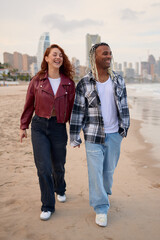 Couple walks hand in hand on the beach during sunset in a cityscape