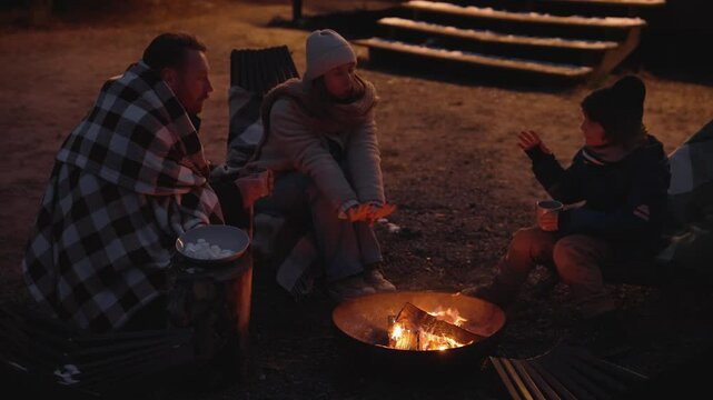 A white adult man and woman sit with a school-aged boy around a campfire, drinking from metal mugs. They sit on chairs on gravel in front of wooden steps with a bowl of marshmallows beside them.