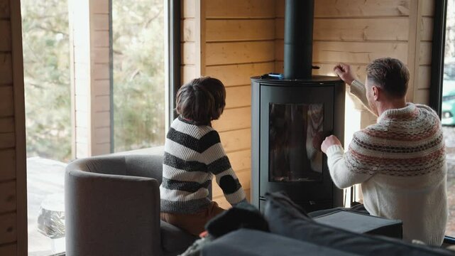 A white adult man and a school-aged boy light a black fireplace inside a wooden cabin. They wear patterned sweaters and sit near large windows on a winter day.