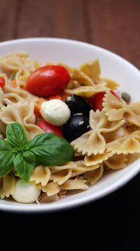 Video verticale, piatto bianco che ruota con pasta fredda condita con pomodori, olive, mozzarelle, foglie di basilico