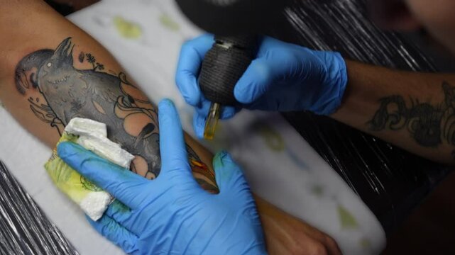 Close-up view of a tattoo artist in blue gloves creating a floral tattoo on a clients forearm. The tattoo machine is in motion as the artist outlines the design. Studio environment