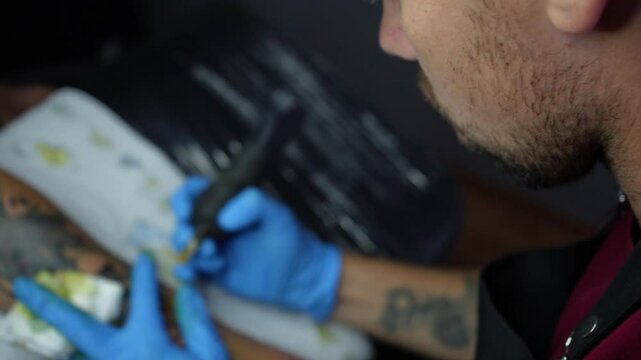 Top-down view of a tattoo artist using a tattoo machine during a session. The artist is focused on the process, wearing gloves. Tattooing in progress. 