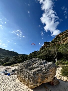 Large stone, boulder on a picturesque beach. Bright, clear sky, Green mountains and a man with a red parachute, paraplane in the background. Space, nature concept, retreat, sport, hobby, recreation