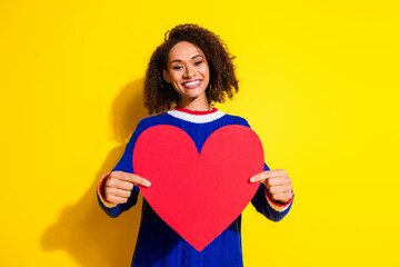 Smiling young woman holding a red heart shape on a vibrant yellow background, expressing joy and...