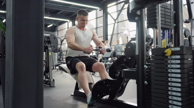 Muscular man performs seated cable row on low row machine pulling heavy weight with full strength in busy modern gym environment while towel draped on bench and other gym goers move around
