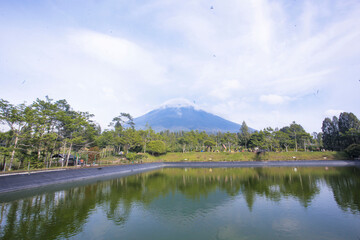 Obraz premium Volcano Reflection: A serene pond reflects the majestic peak of a snow-capped volcano, surrounded by lush greenery under a tranquil sky.