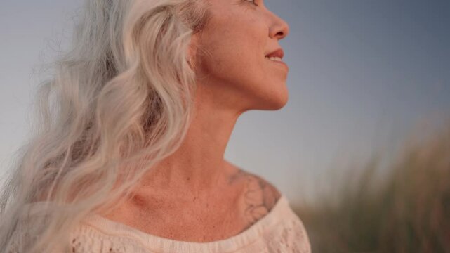 An elderly woman with long gray hair and a tattoo enjoys a calm moment at the beach, embracing peace and tranquility.